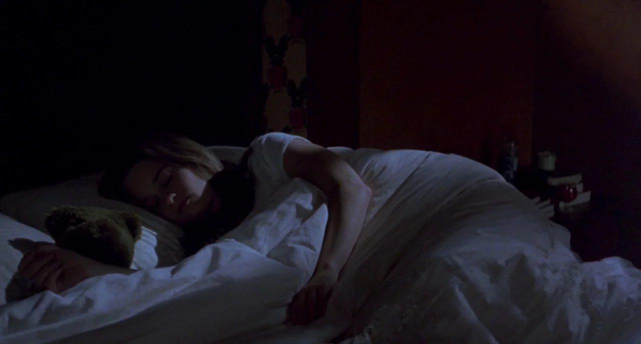 The Craft (1996) movie still: medium — A dark bedroom with patterned wallpaper is dimly lit by a soft light source. A woman sleeps under white…
