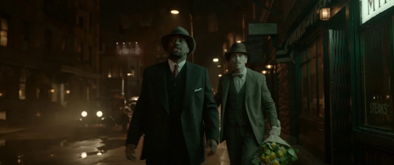 Amsterdam (2022) movie still: fullbody — A wet urban street at night with brick buildings and a Coca-Cola sign. Two men dressed in 1930s style…