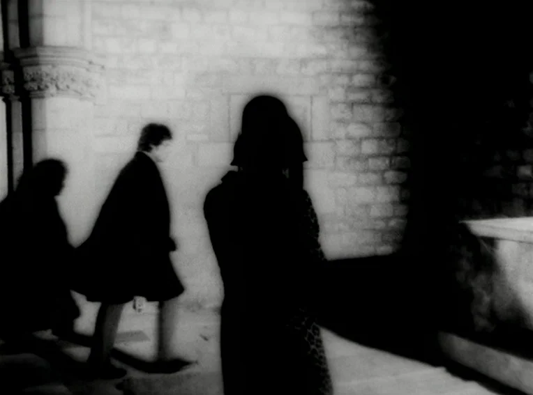 Cuadecuc, Vampir (1971) movie still: medium — A stone-walled interior with blurred figures walking across the frame. Two individuals in dark coats…