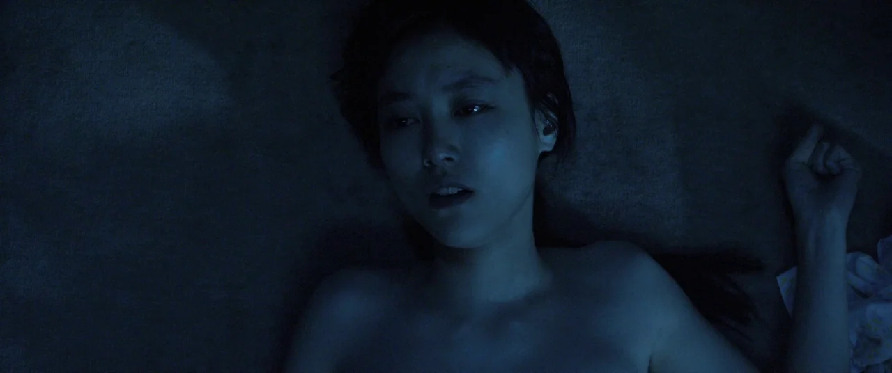 Norwegian Wood (2010) movie still: closeup — A dark, dimly lit indoor setting provides a minimalist backdrop for the subject. A woman lies flat…