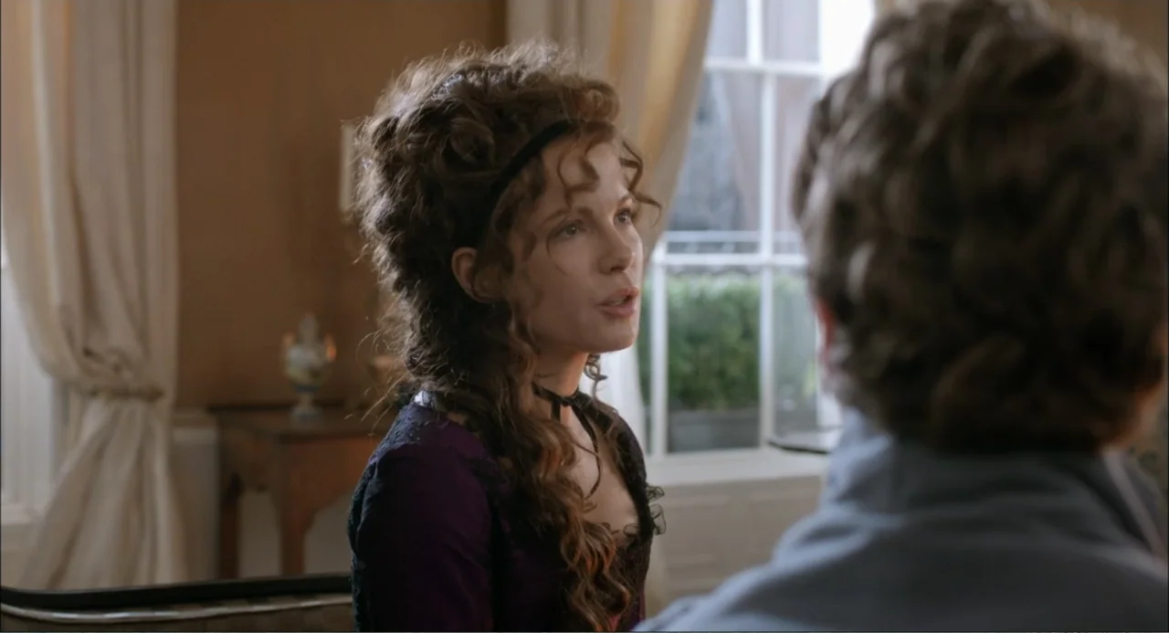Love & Friendship (2016) movie still: medium — A 19th-century parlor with light-colored curtains and a wooden side table visible. A woman with an…