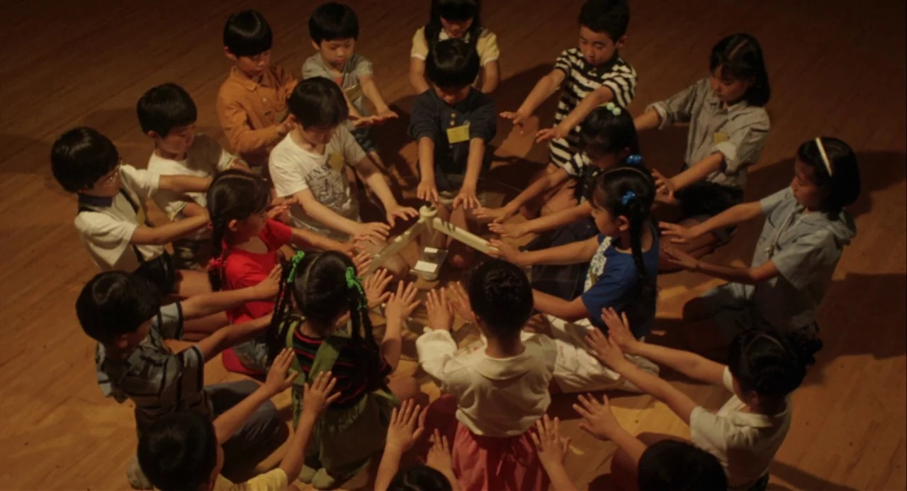 Godzilla vs. Mechagodzilla II (1993) movie still: wide — A group of children sit on a light-colored wooden floor in a circular formation. The…