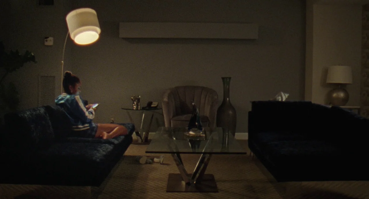 Zola (2020) movie still: wide — A sparsely furnished hotel room features two dark sofas flanking a glass coffee table. A young woman sits alone on…
