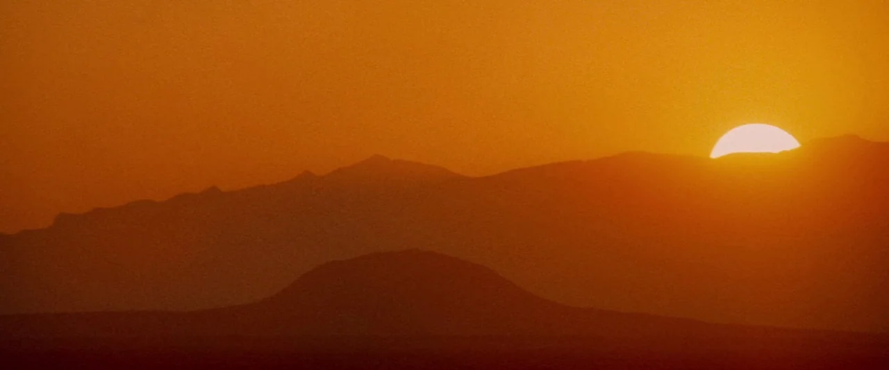 Conan The Barbarian (1982) movie still: establishing — A vast, arid landscape features layered, shadowed mountain silhouettes against a bright…