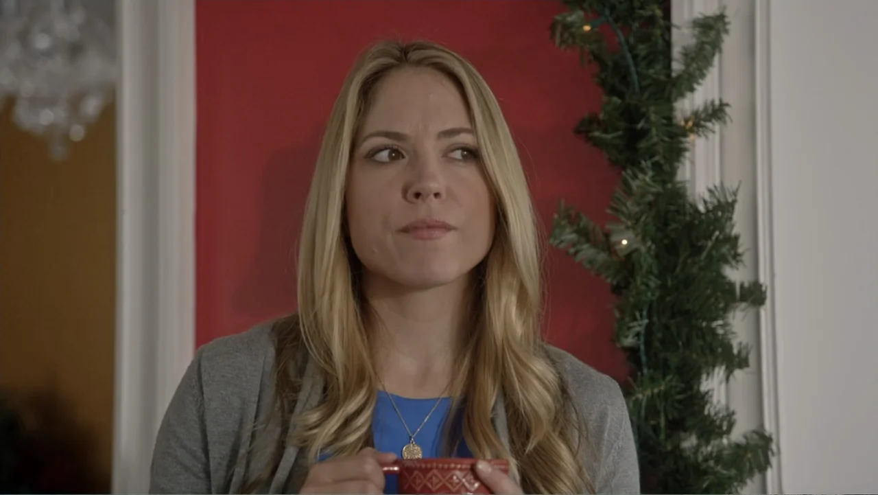 The Christmas Cure (2017) movie still: medium — A woman stands indoors in front of a red wall adorned with Christmas garland. She holds a red…