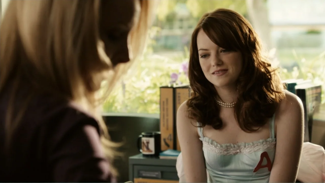 Easy A (2010) movie still: medium — A high school administrative office featuring binders on a desk and bright window light. A young woman in a pale…
