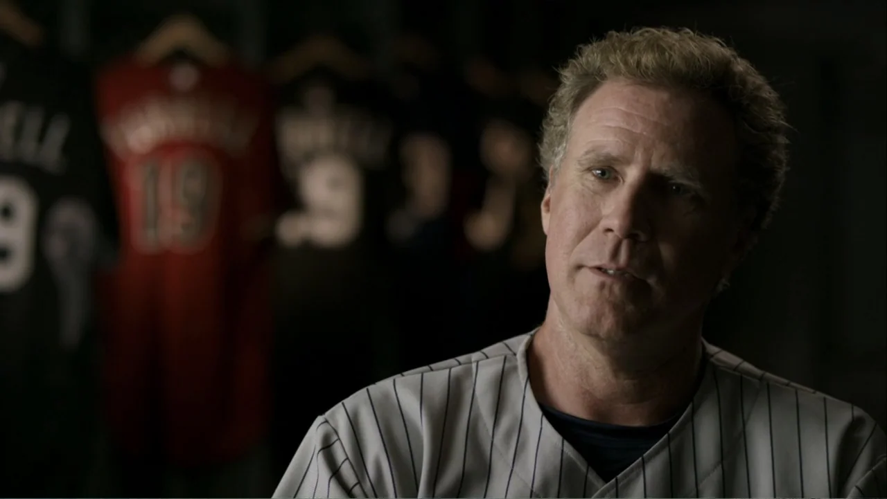 Ferrell Takes the Field (2015) movie still: medium — A dimly lit locker room features rows of hanging baseball jerseys in the background. A…