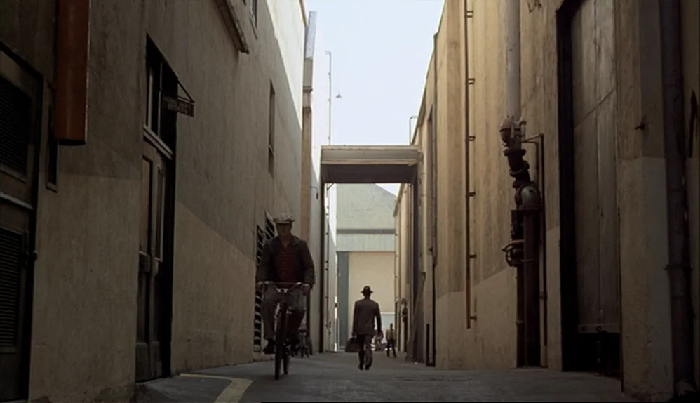 The Godfather (1972) movie still: wide — A narrow alleyway with tall beige concrete walls and industrial metal pipework. A man rides a bicycle…