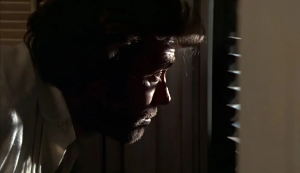 The Beguiled (1971) movie still: closeup — An interior space features horizontal blinds filtering light into a darkened room. A man with stubble…
