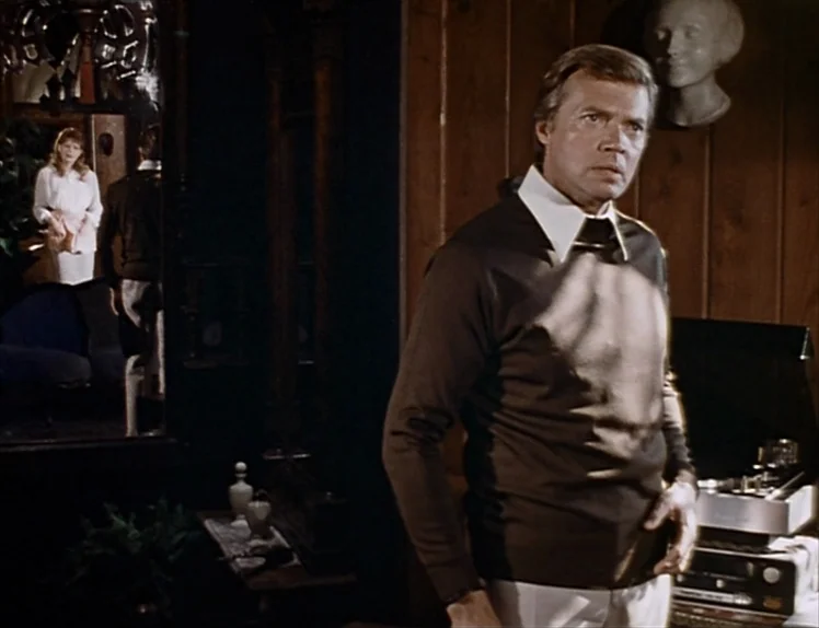 Martha (1974) movie still: medium — A dimly lit room features wood paneling and a reflective surface on the left. A middle-aged man stands in the…