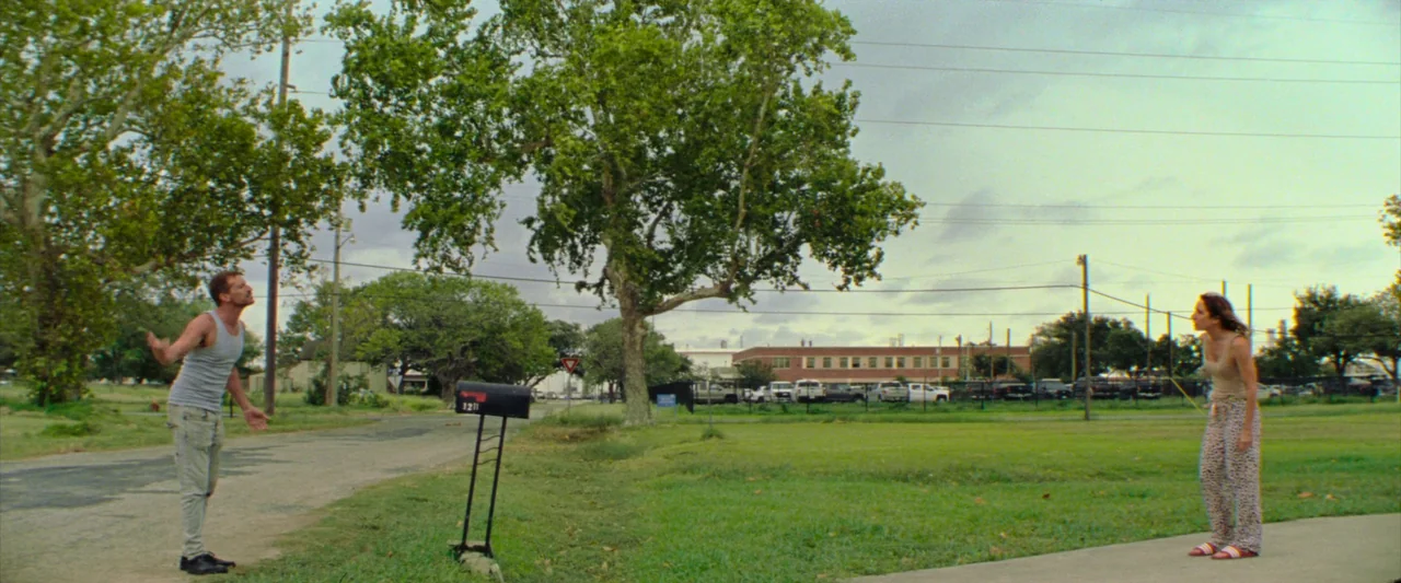 Red Rocket (2021) movie still: wide — A residential street with a grassy yard and large oak trees under overcast sky. A man stands on the road while…