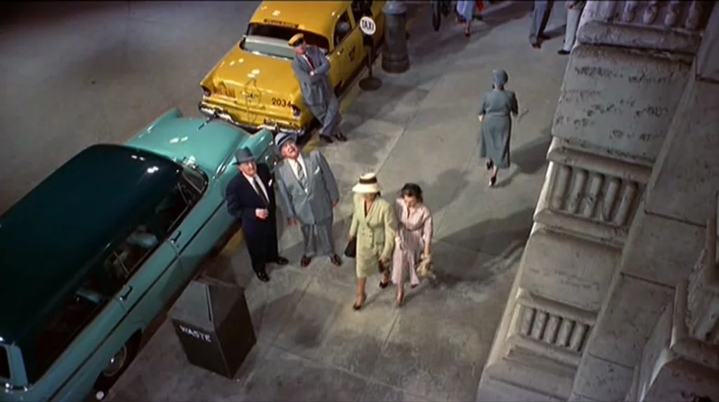 Artists and Models (1955) movie still: wide — An urban sidewalk outside a large building features vintage automobiles and pedestrians at night. A…