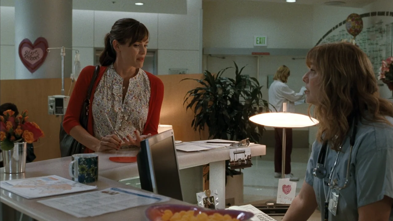 F Valentine's Day (2026) movie still: medium — A hospital reception area features a desk with papers, a plant, and a desk lamp. A woman in a red…