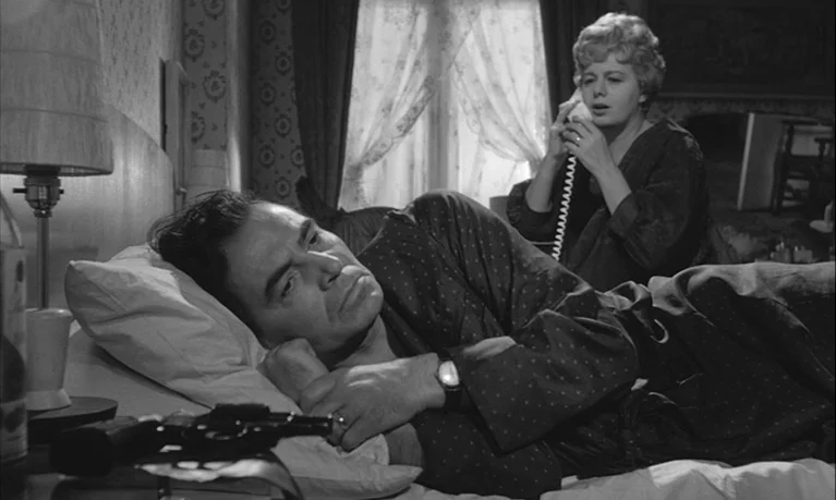 Lolita (1962) movie still: medium — A hotel room with patterned wallpaper and a window with lace curtains. A man lies on the bed in the foreground…