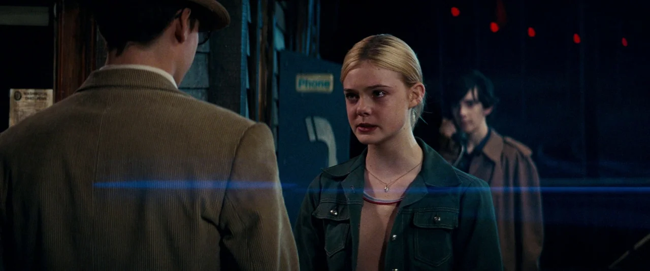 Super 8 (2011) movie still: medium — A night setting outdoors with a payphone stall visible behind a blonde girl. The girl stands facing a boy while…