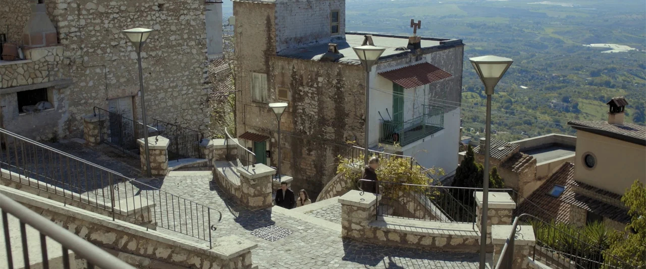About Us (2017) movie still: wide — A historic stone village perched on a hill features narrow cobblestone walkways and stairs. Two men and a woman…