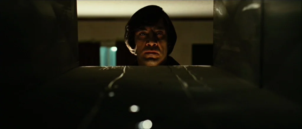 No Country For Old Men movie still: closeup — A man looks directly into the lens from behind a narrow vent opening. The metal surface of the air…