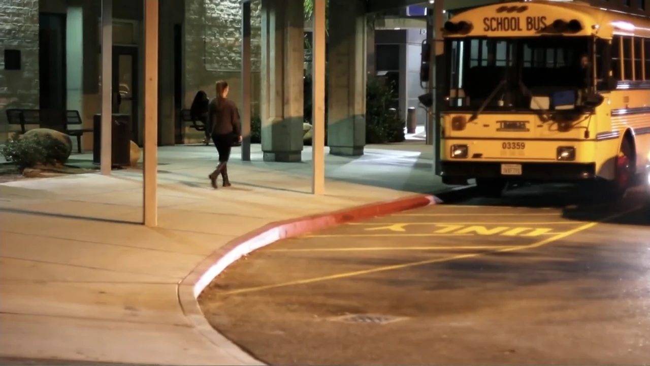 The (Dead Mothers) Club (2014) movie still: medium — A nighttime school bus loading zone features a concrete overhang and a yellow bus. A woman…