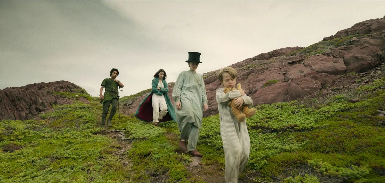 Peter Pan & Wendy (2023) movie still: fullbody — A group of children in pajamas and costumes traverse a barren rocky, moss-covered hill. The…