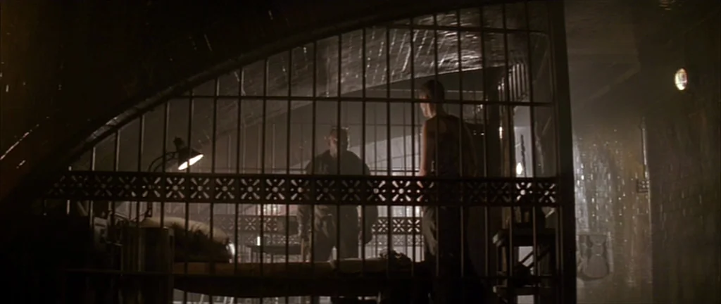 Alien 3 (1992) movie still: wide — An interior space enclosed by heavy iron bars and dark brick walls. Two men stand together inside the cage…