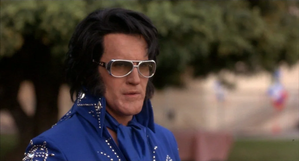 Bubba Ho-Tep (2002) movie still: medium — A man wearing an Elvis Presley costume stands outside in a public park setting. The background features…