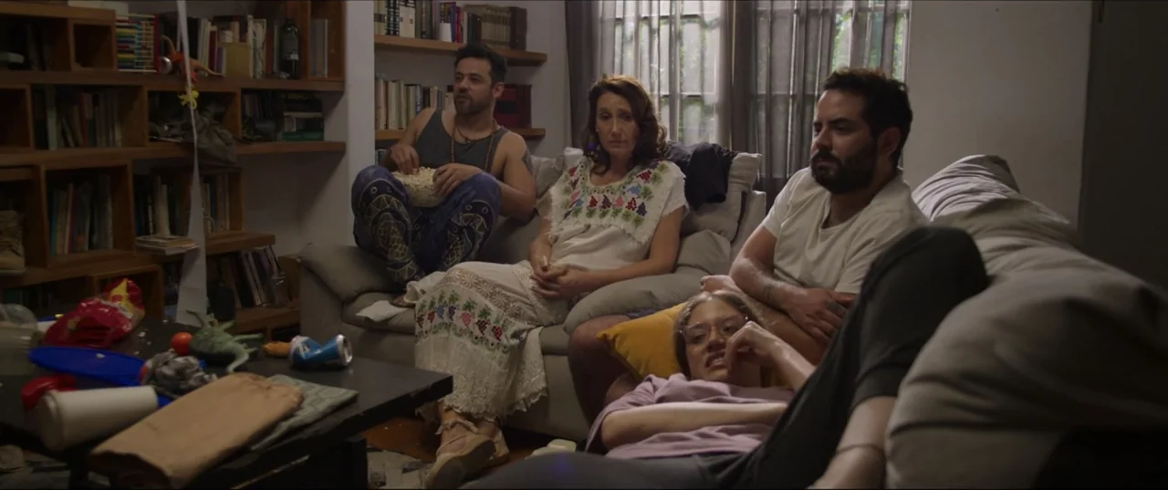 Can't Quit Mom (2022) movie still: medium — A group of four adults sit together on a sofa in a cluttered room. They watch a screen while one person…
