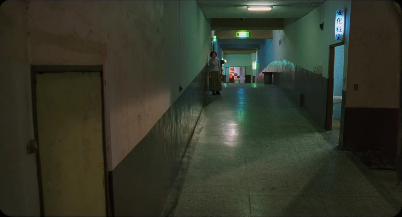 Goodbye, Dragon Inn (2003) movie still: wide — A long, narrow institutional hallway features mottled concrete floors and plain, painted walls. A…