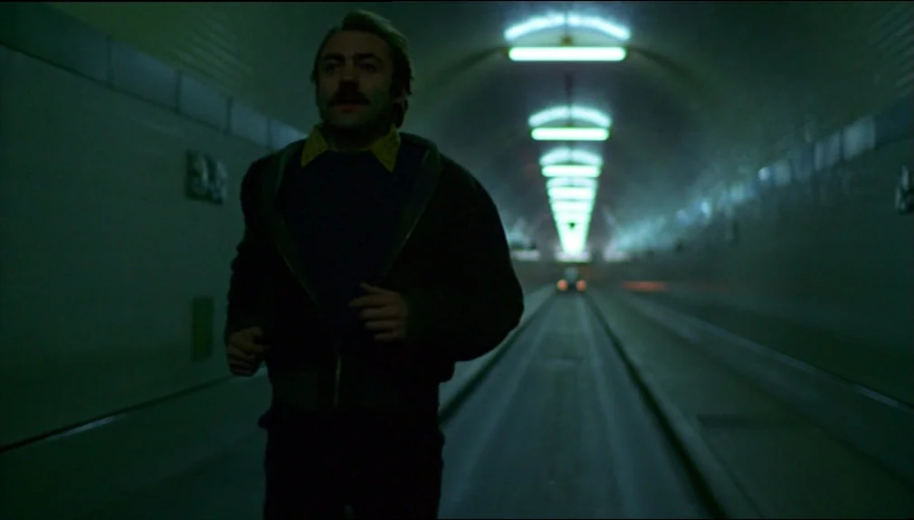 The American Friend (1977) movie still: medium — A long, curved concrete tunnel illuminated by a series of receding overhead fluorescent lamps. A…