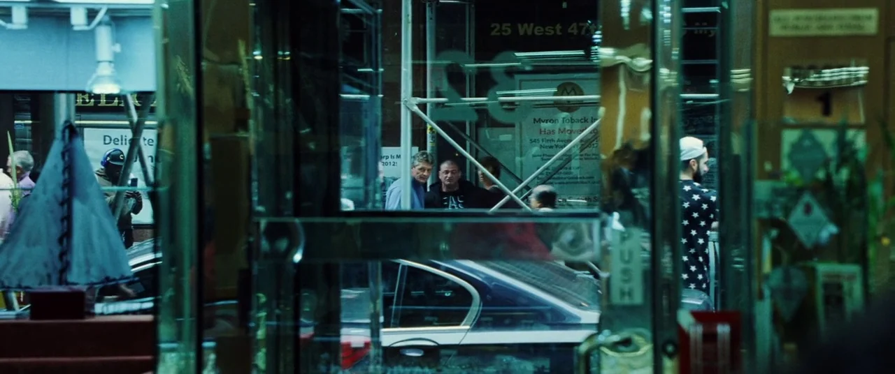 Uncut Gems (2019) movie still: wide — A view through a glass entryway looking out onto a dark New York street. People move past scaffolding in front…