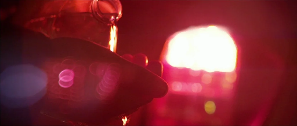 Kill List movie still: closeup — A dark, blurred interior space is illuminated by a singular, intense glowing red light source. A clear liquid pours…