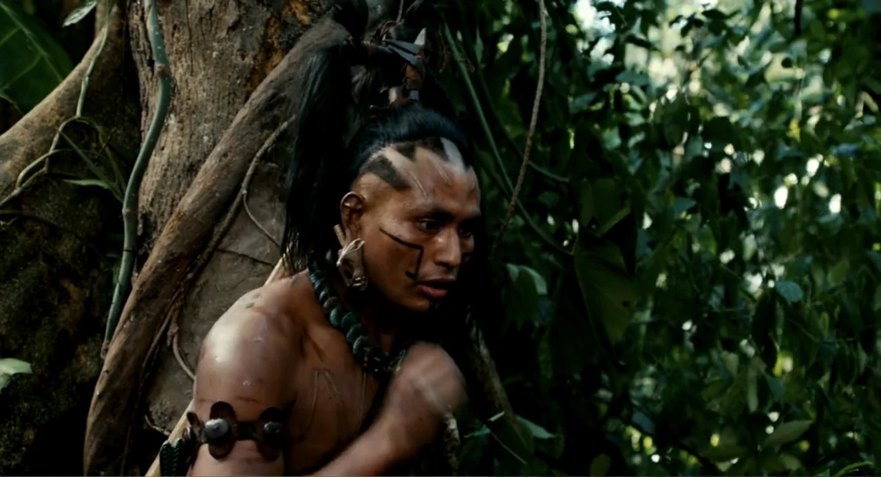 Apocalypto (2006) movie still: medium — A dense jungle environment with thick vegetation and large tree trunks visible behind. A man with elaborate…