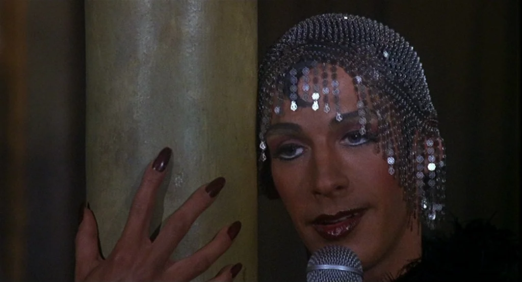 Deux (2002) movie still: closeup — A dimly lit club interior featuring a structural column and a stage area. A person wears a silver sequined…