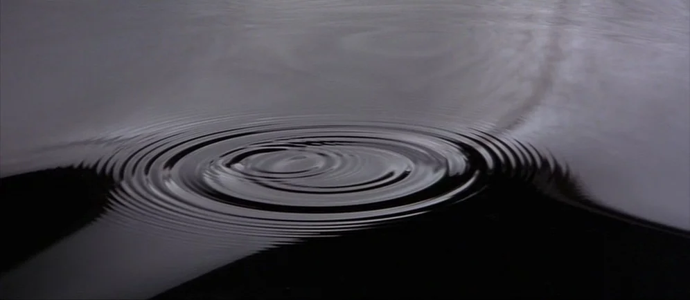 Dune (1984) movie still: closeup — A dark, calm body of water fills the frame with subtle light reflections. Ripples radiate outwards from a central…