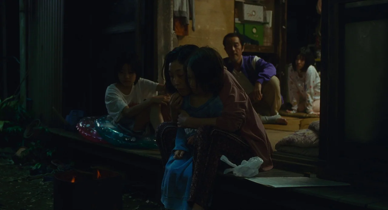 Shoplifters (2018) movie still: medium — A group of people sit on the wooden veranda of a traditional house at night. A small fire burns in a metal…