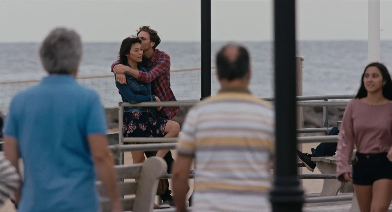 Lingua Franca (2019) movie still: wide — A public boardwalk overlooking an ocean horizon under an overcast sky. A couple embraces behind a metal…