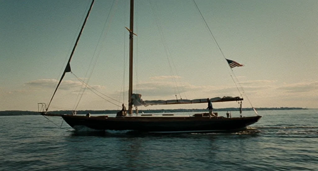Burn After Reading (2008) movie still: wide — A classic wooden sailboat glides across calm water under a clear sky. A man walks across the deck…