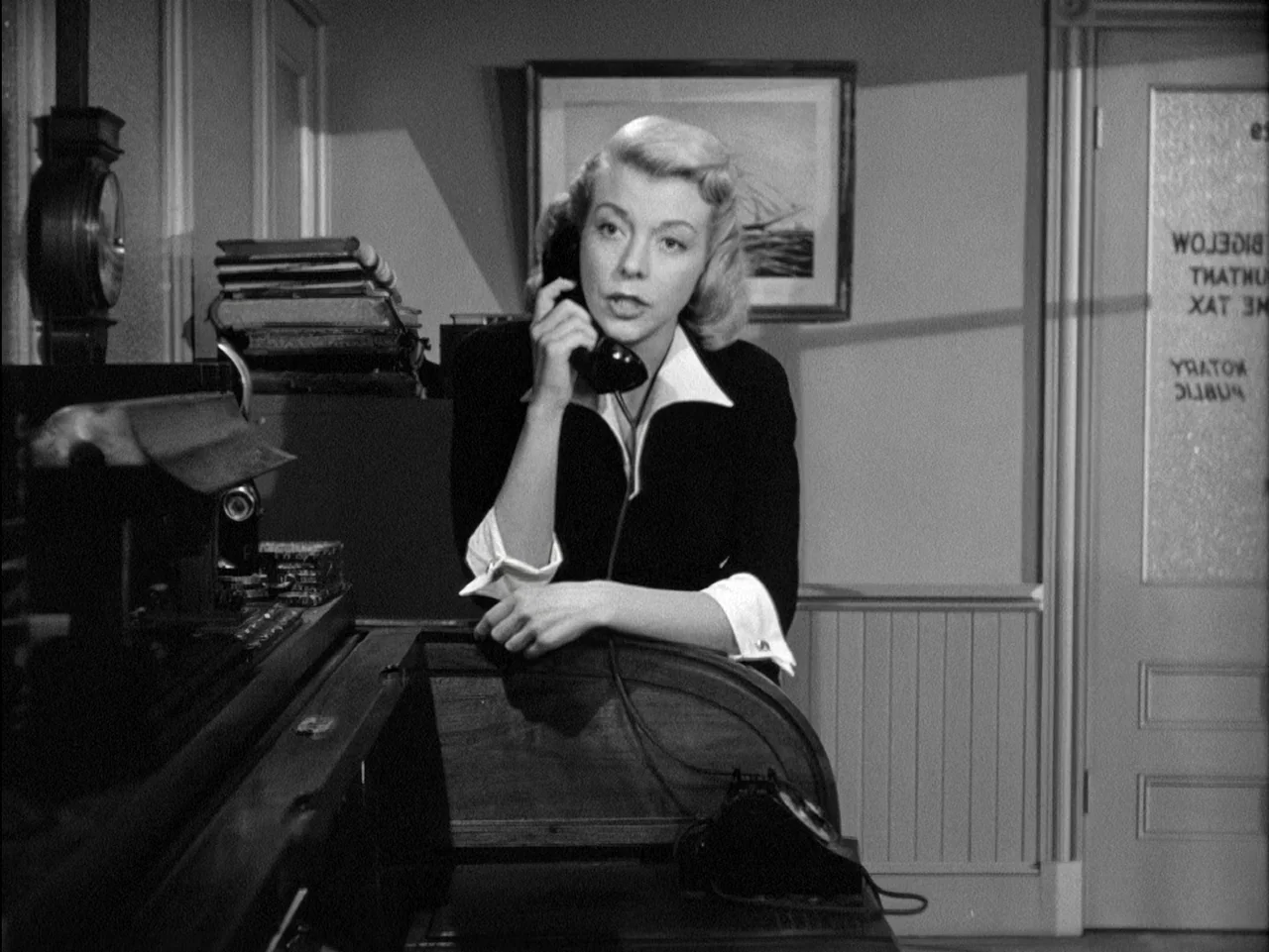 D.O.A. (1949) movie still: medium — An office features a wooden desk with a stack of books and a telephone. A blonde woman in a blazer stands behind…