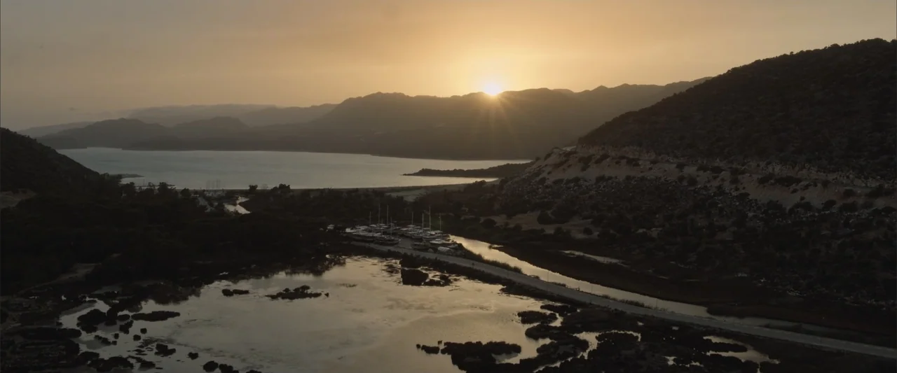 Blue Cave (2024) movie still: establishing — A wide coastal inlet is surrounded by low mountains during the late afternoon. Several small boats are…