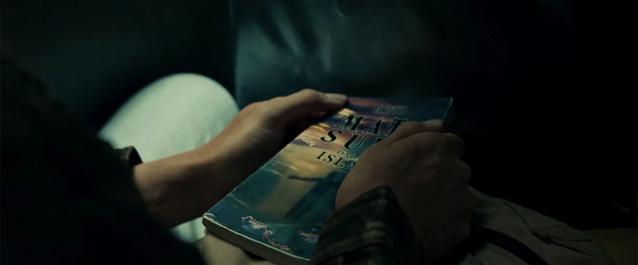 The Unholy (2022) movie still: closeup — A person holds a paperback book in their lap within a dark vehicle. The book cover features an image of a…