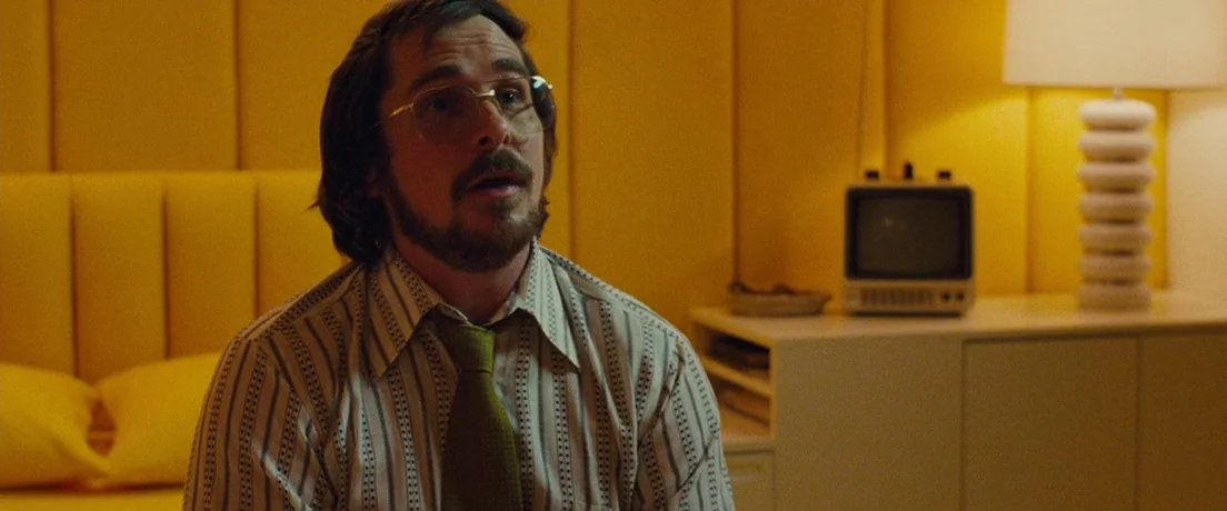 American Hustle (2013) movie still: medium — A 1970s hotel room features a bright yellow upholstered headboard and matching lighting. A man with a…