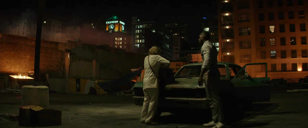 Hotel Artemis (2018) movie still: wide — A dark urban parking lot at night features a teal vintage sedan parked centrally. The man and woman stand…