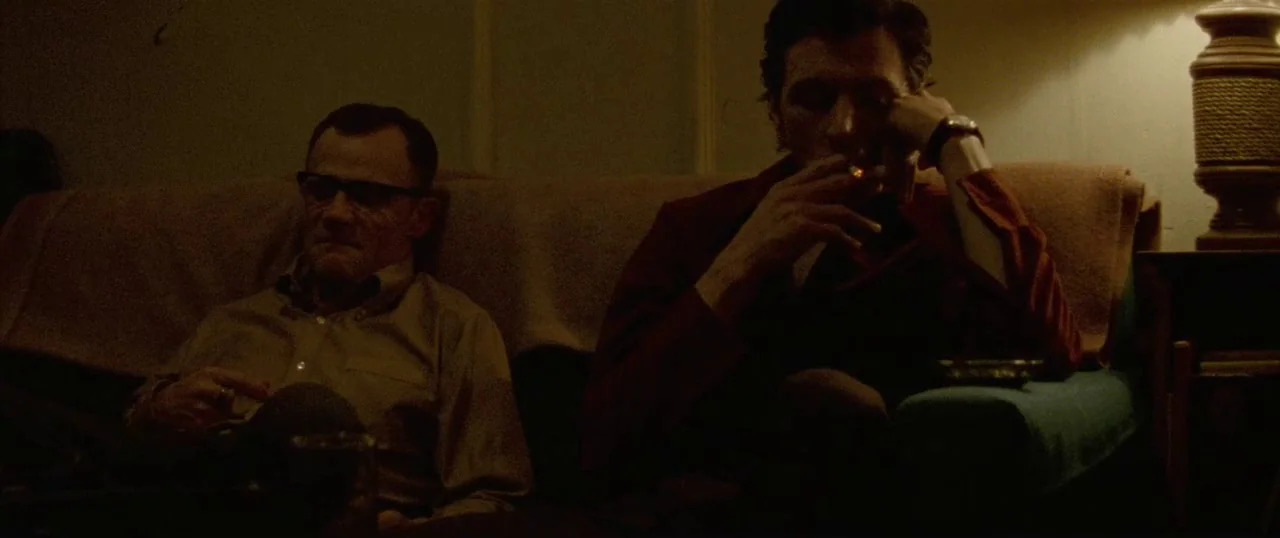 Low Down (2014) movie still: medium — A dimly lit room with mid-century furniture and muted brown wall tones. Two men sit on a couch in silence…