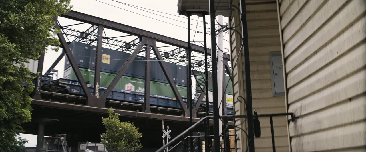 Dogman (2023) movie still: wide — A rusted iron railway bridge spans across the upper frame over a suburban street. A freight train with…