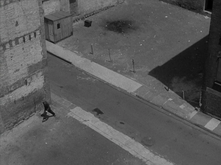 Shadow of a Doubt (1943) movie still: wide — An urban lot features a weathered brick wall and a small wooden shed nearby. A lone man moves across…