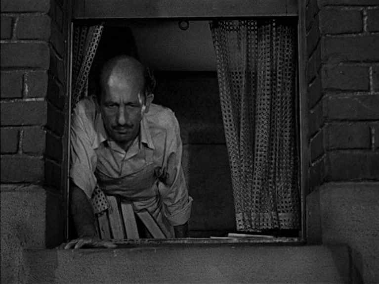 The Big Night (1951) movie still: medium — A man peers out from a small window framed by brick masonry and net curtains. The subject leans forward…