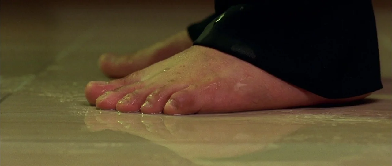 Sympathy for Mr. Vengeance (2002) movie still: closeup — A low-angle view of bare human feet standing on a wet tiled floor. Dark fabric cuffs are…