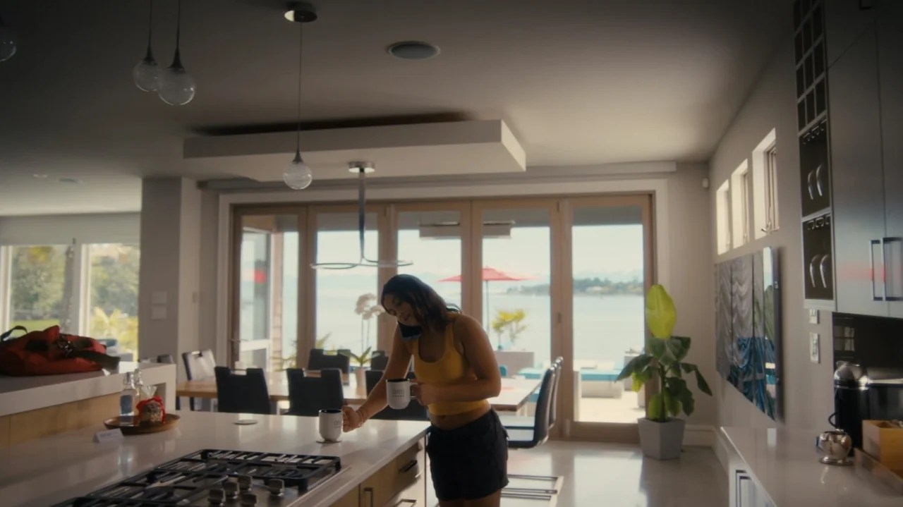 Sidelined: The QB and Me (2025) movie still: medium — A sunlit modern house with a large open floor plan and water views. A young woman stands at…