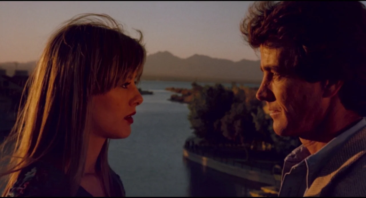 Olivia (1983) movie still: medium — Two individuals stand facing each other on a balcony overlooking a lake at sunset. Distant mountains frame the…