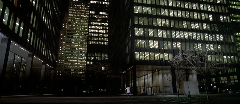 American Psycho (2000) movie still: establishing — A dark, modern urban plaza is framed by towering skyscraper glass facades at night. The windows…