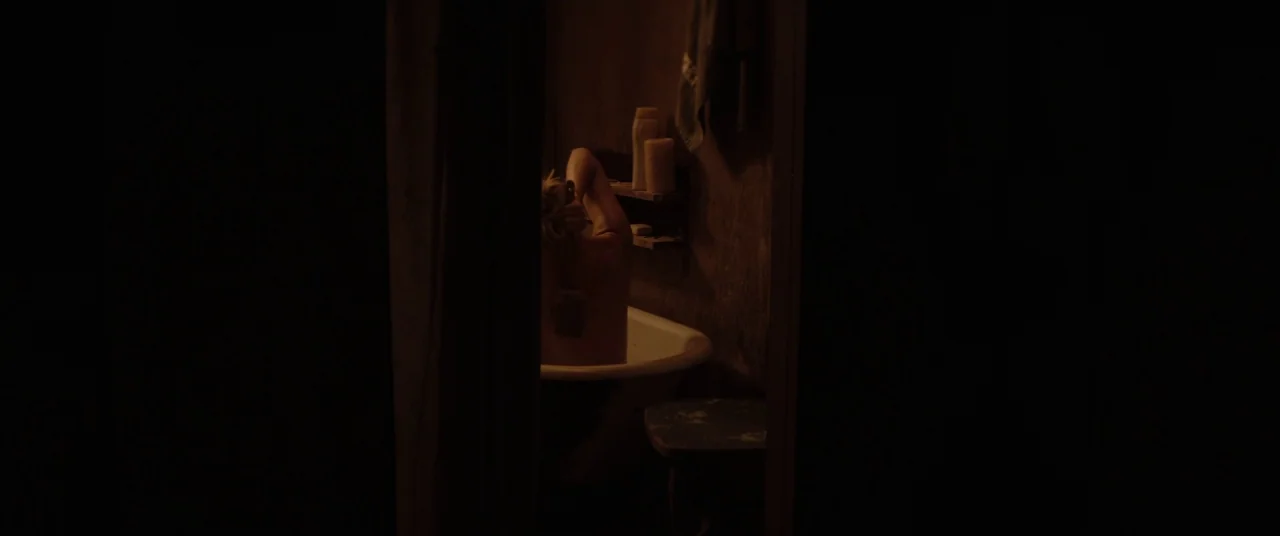 Hold The Dark (2018) movie still: medium — A dimly lit room features an old porcelain bathtub against a dark wall. A woman sits in the tub using a…